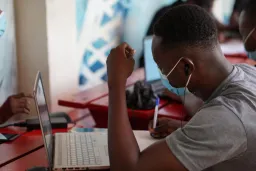 Man writing in notebook in front of laptop, wearing COVID mask