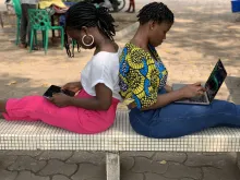 Two women sitting with their backs facing each other. One is on a laptop and the other is on a tablet